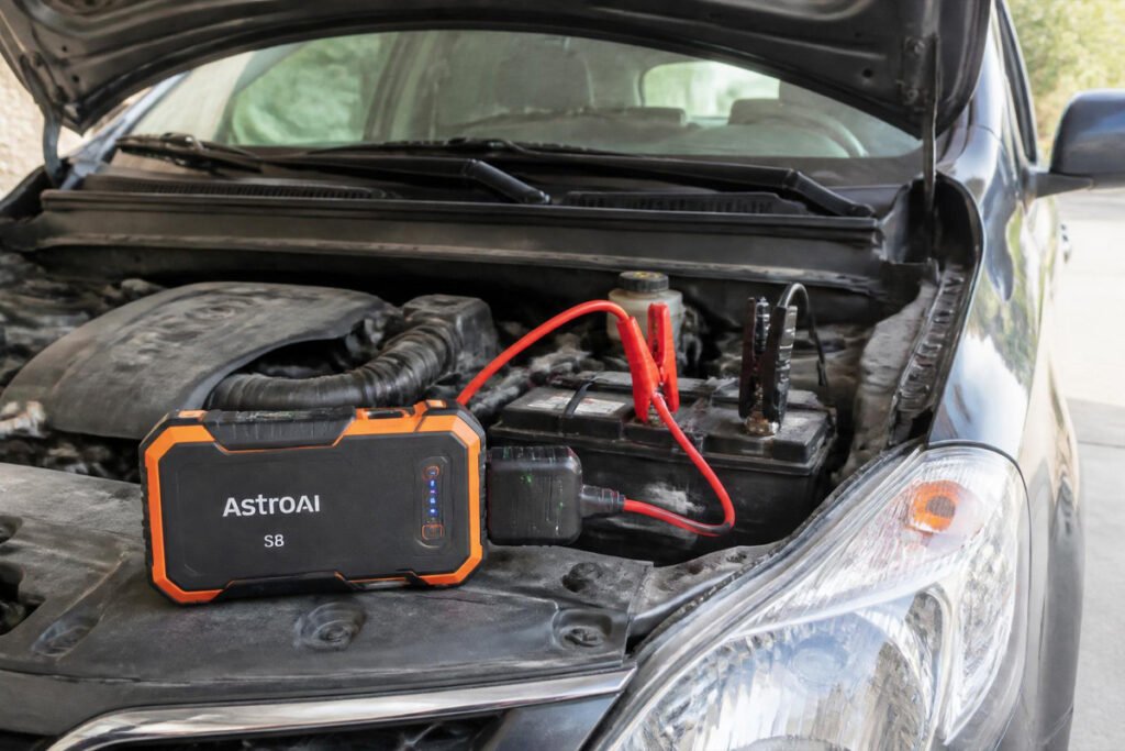 AstroAI S8 compact jump starter next to a small commuter car, clamps visible